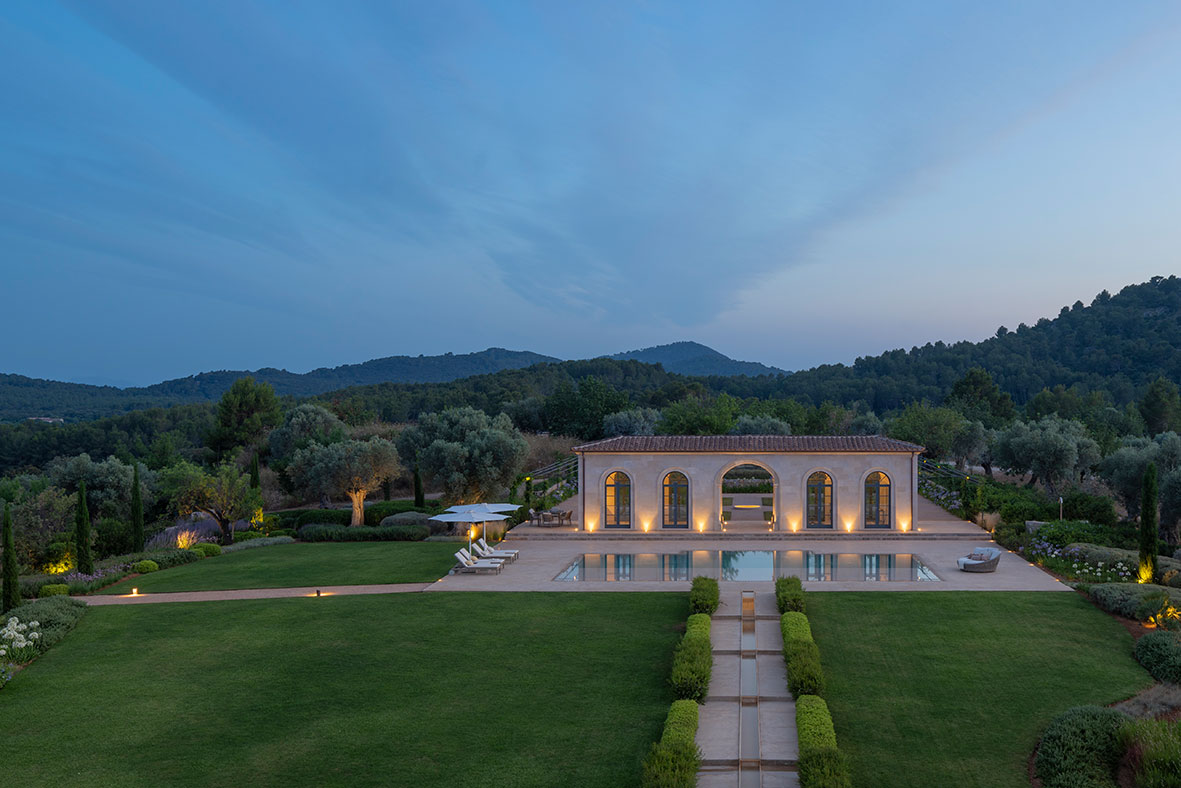 Blick aus erhöhter Perspektive auf den beleuchteten Pavillon und den Pool am Abend, mit Rasen, Wasserkanal und Bergen
