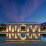 Pavillon mit beleuchteten Bögen und großen Fenstern am Abend, im Pool gespiegelt unter tiefblauem Himmel