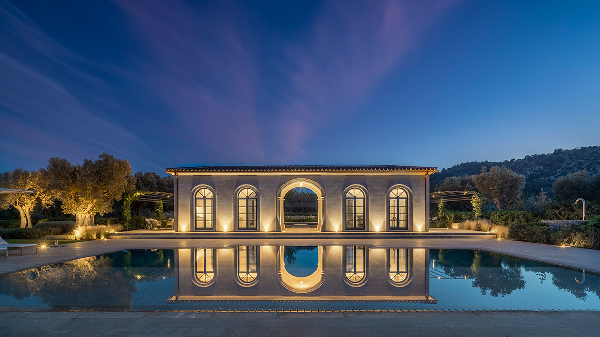 Pavillon mit beleuchteten Bögen und großen Fenstern am Abend, im Pool gespiegelt unter tiefblauem Himmel