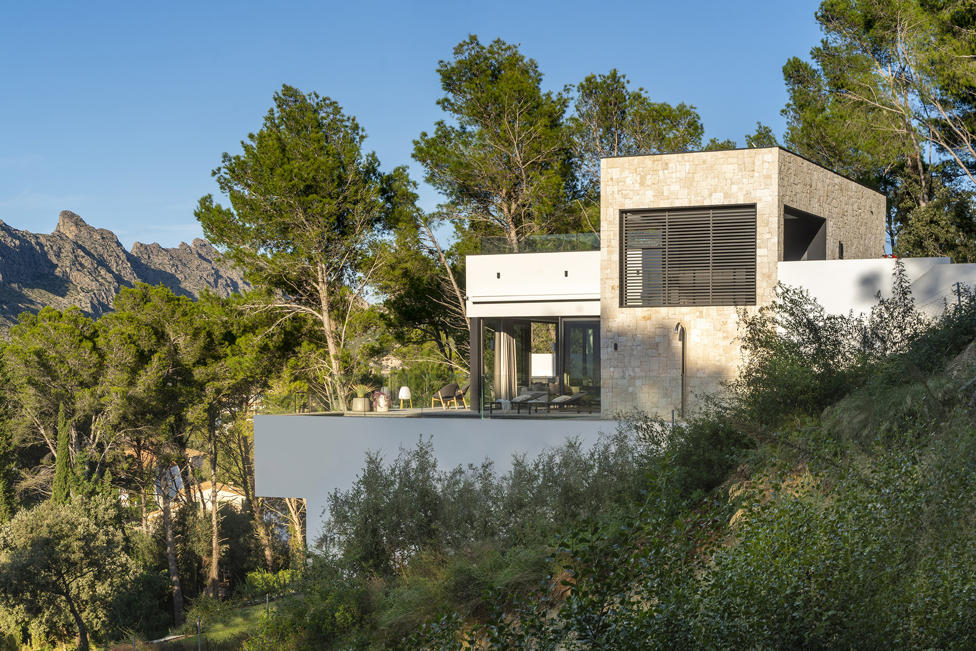 Fassade aus hellem Stein und weißen Mauern mit auskragender Terrasse und Bergen im Hintergrund