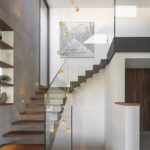 Treppe mit frei auskragenden Holzstufen, Glasgeländer, bernsteinfarbenen Hängeleuchten und Regalen an grauer Wand
