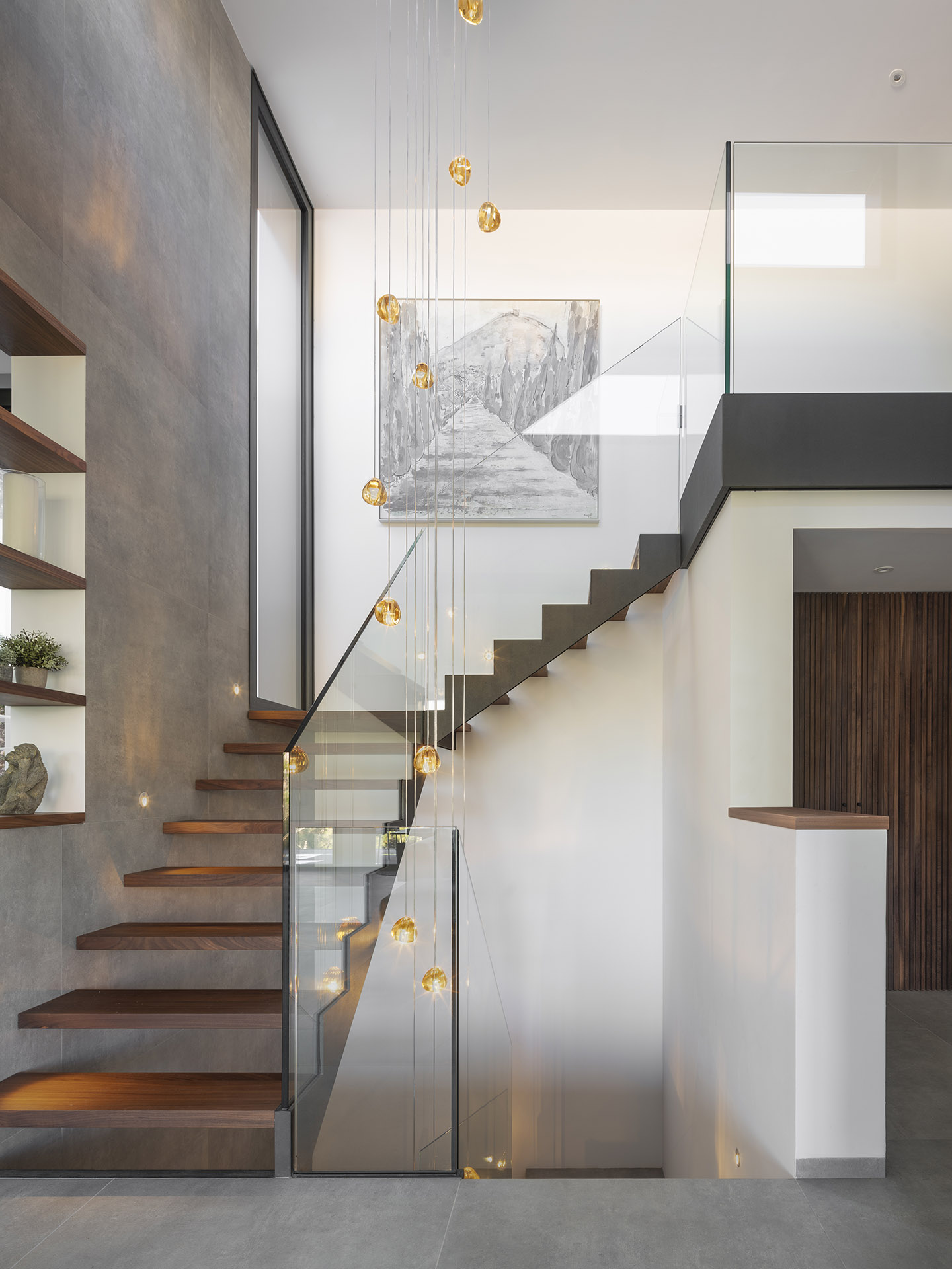 Treppe mit frei auskragenden Holzstufen, Glasgeländer, bernsteinfarbenen Hängeleuchten und Regalen an grauer Wand