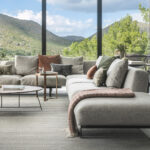 Graues Modulsofa mit terrakottafarbenen und grünen Kissen vor großen Fenstern mit Blick auf die Berge