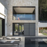 Fassade aus Naturstein mit verglaster oberer Terrasse, Sonnenliegen und Swimmingpool mit dunklem Mosaik