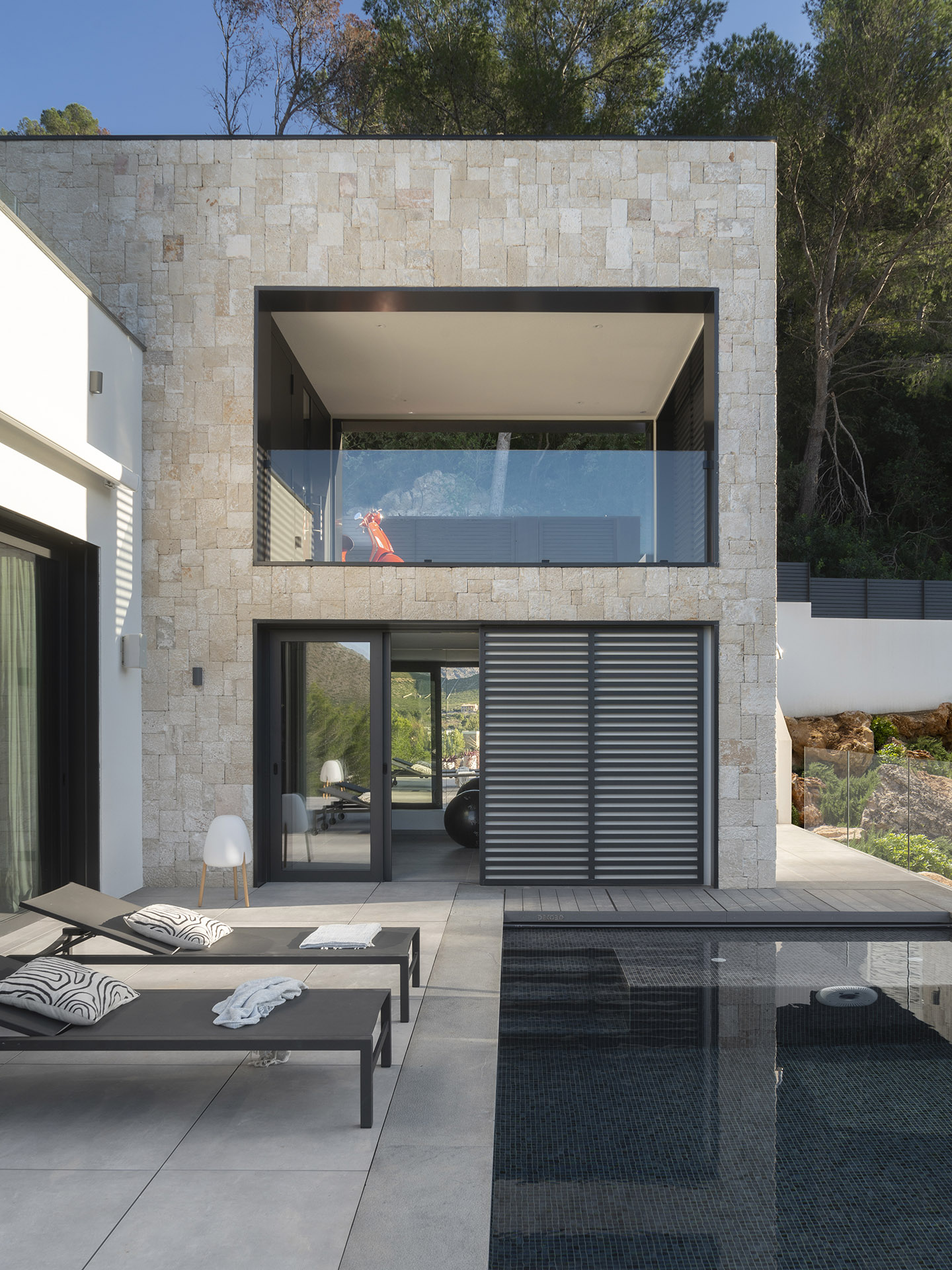 Fassade aus Naturstein mit verglaster oberer Terrasse, Sonnenliegen und Swimmingpool mit dunklem Mosaik