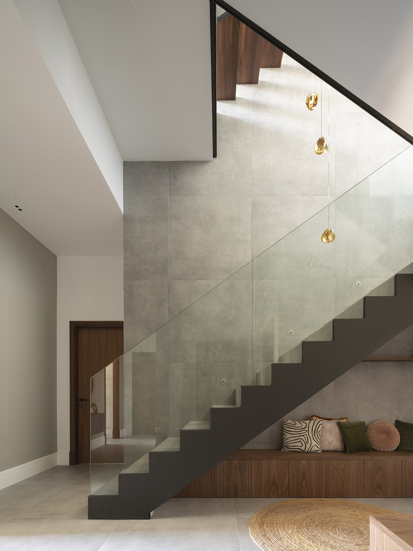 Dunkle Treppe mit Glasgeländer, Wand aus grauem Feinsteinzeug, bernsteinfarbenen Hängeleuchten und Holzbank