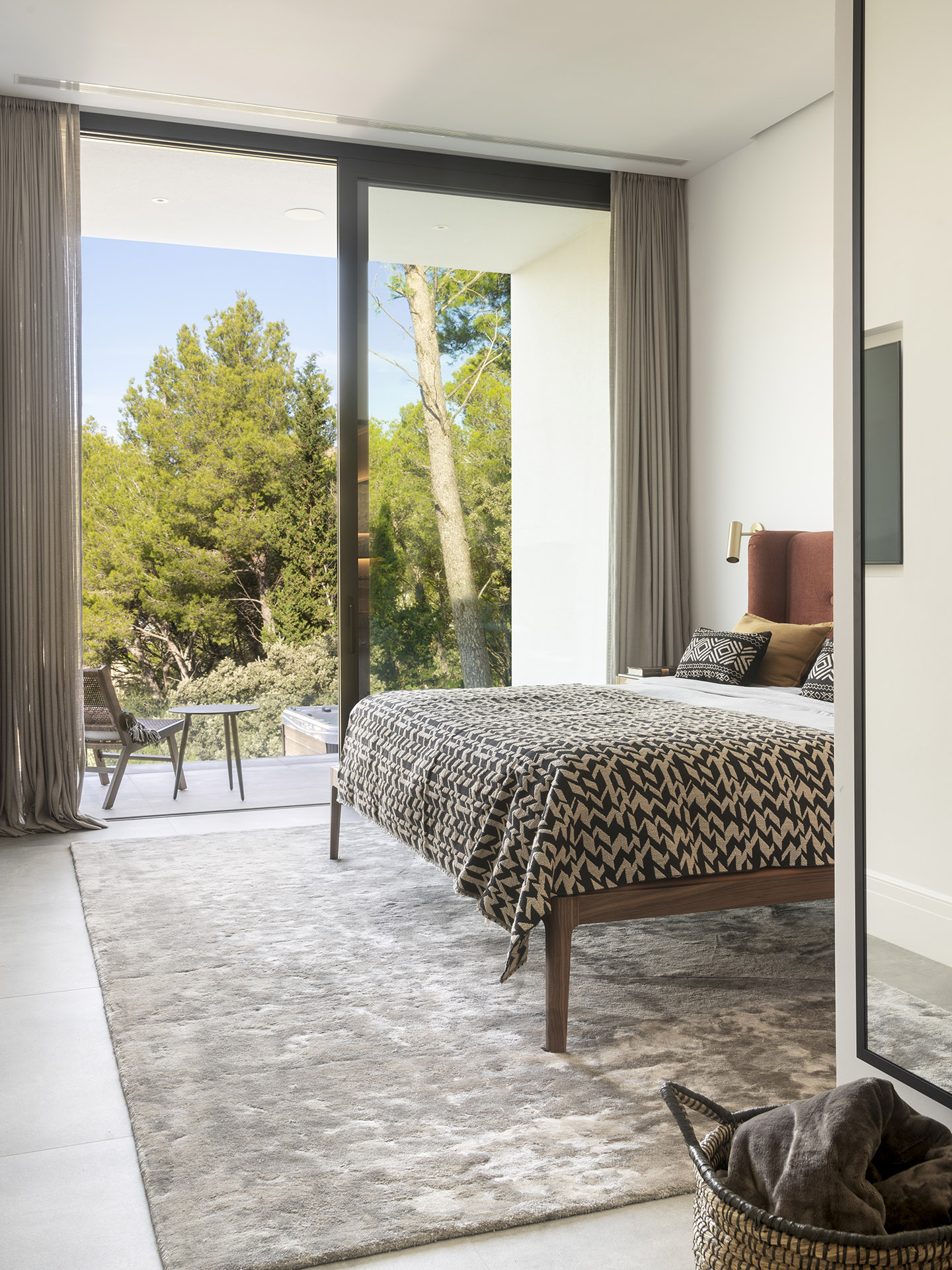 Schlafzimmer mit Holzbett, schwarz-beige gemusterter Tagesdecke und großem Fenster zum Kiefernwald