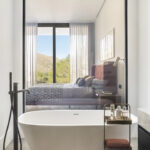 Freistehende weiße Badewanne neben einer Glaswand mit Durchblick auf das Schlafzimmer und die Berge