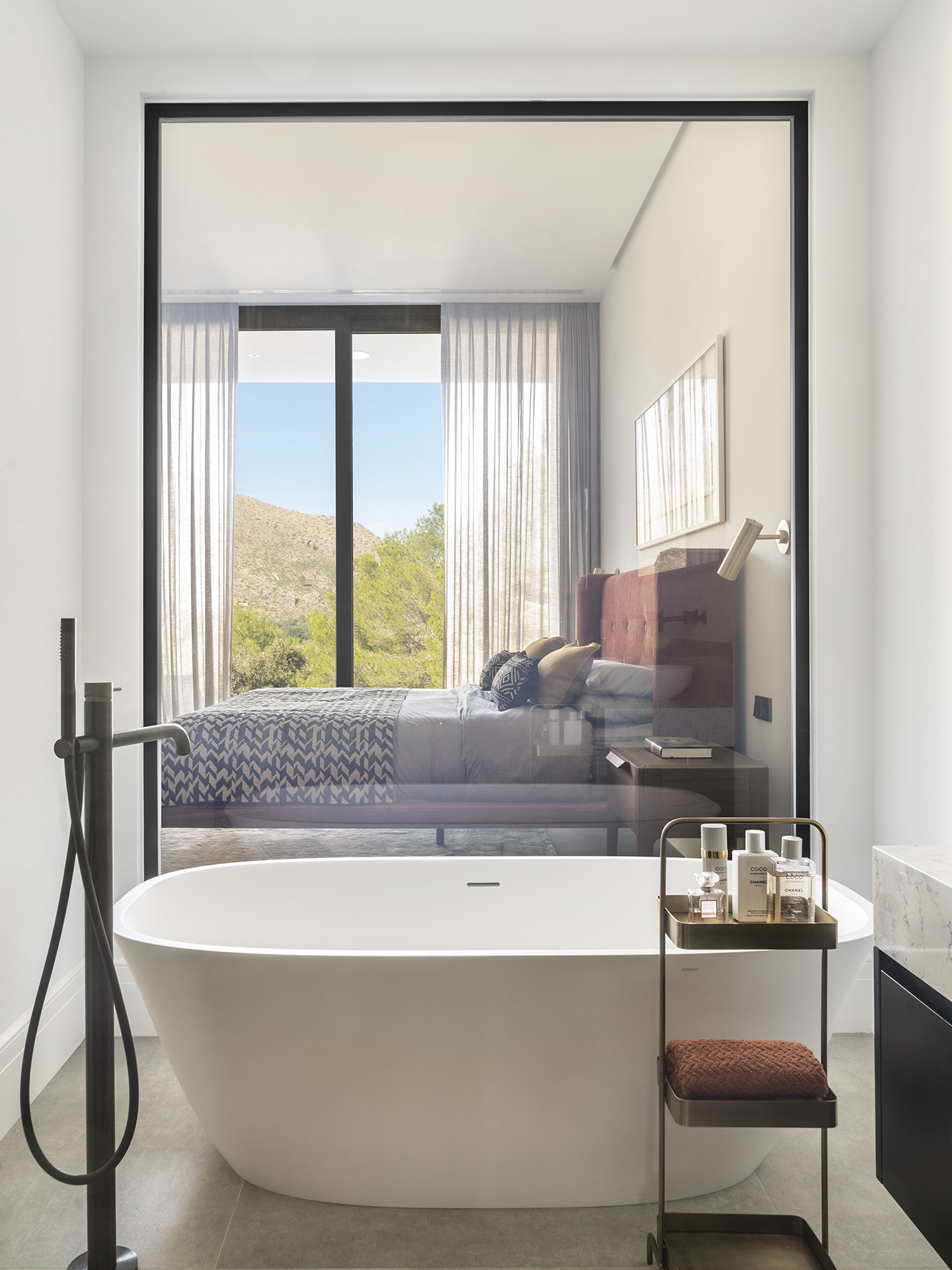 Freistehende weiße Badewanne neben einer Glaswand mit Durchblick auf das Schlafzimmer und die Berge