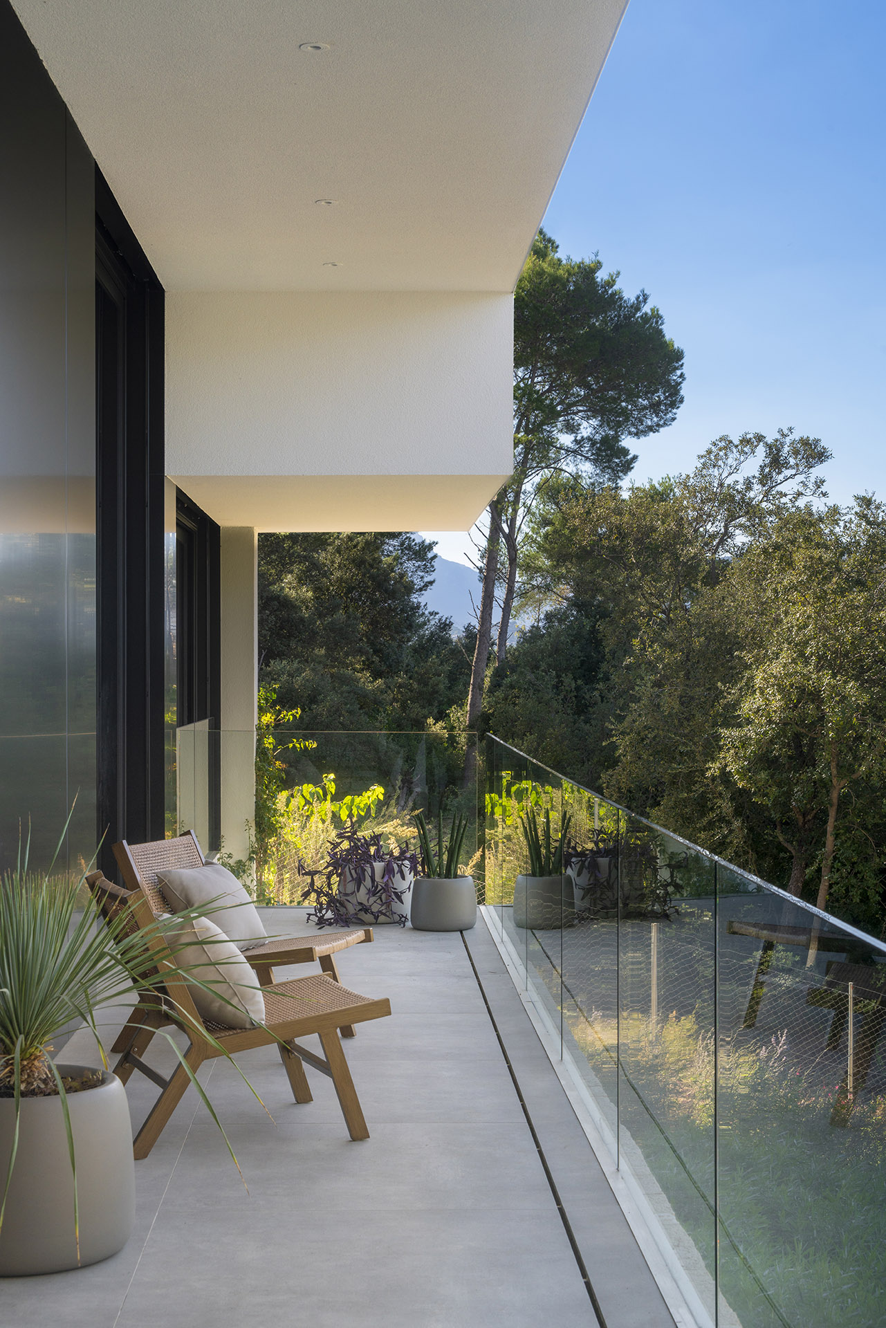 Terrasse mit Sesseln aus Holz und Geflecht, Blumentöpfen und Glasgeländer, offen zum Kiefernwald