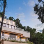 Fassade in der Abenddämmerung mit gestaffelten Terrassen, Glasgeländern und warmer Beleuchtung zwischen Kiefern