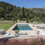 Blick von oben auf den Swimmingpool, umgeben von Liegen, gemauerten Sofas, Steinmauer und rosa Blüten