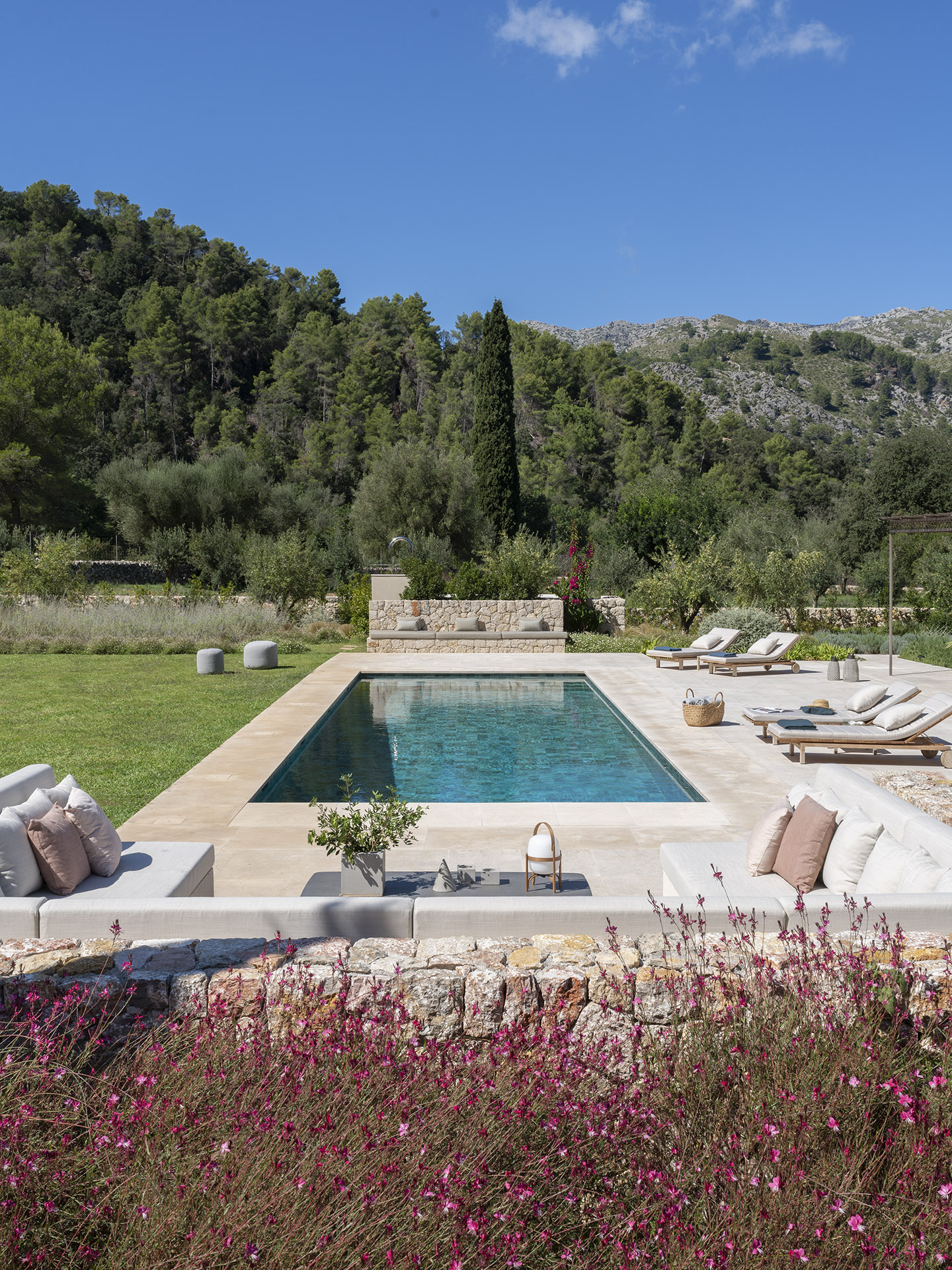 Blick von oben auf den Swimmingpool, umgeben von Liegen, gemauerten Sofas, Steinmauer und rosa Blüten