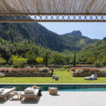 Blick unter der Pergola aus Schilfrohr auf den Swimmingpool, die Sonnenliegen, den Garten mit Steinterrassen und den Berg