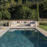 Frontalansicht des Swimmingpools mit gemauertem Sofa im Hintergrund, Liegen links und Poufs auf dem Rasen