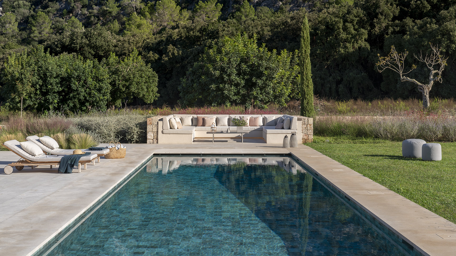 Frontalansicht des Swimmingpools mit gemauertem Sofa im Hintergrund, Liegen links und Poufs auf dem Rasen