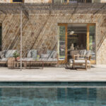 Terrasse am Swimmingpool mit Holzsofas, Pergola aus Schilfrohr und Steinfassade im Hintergrund