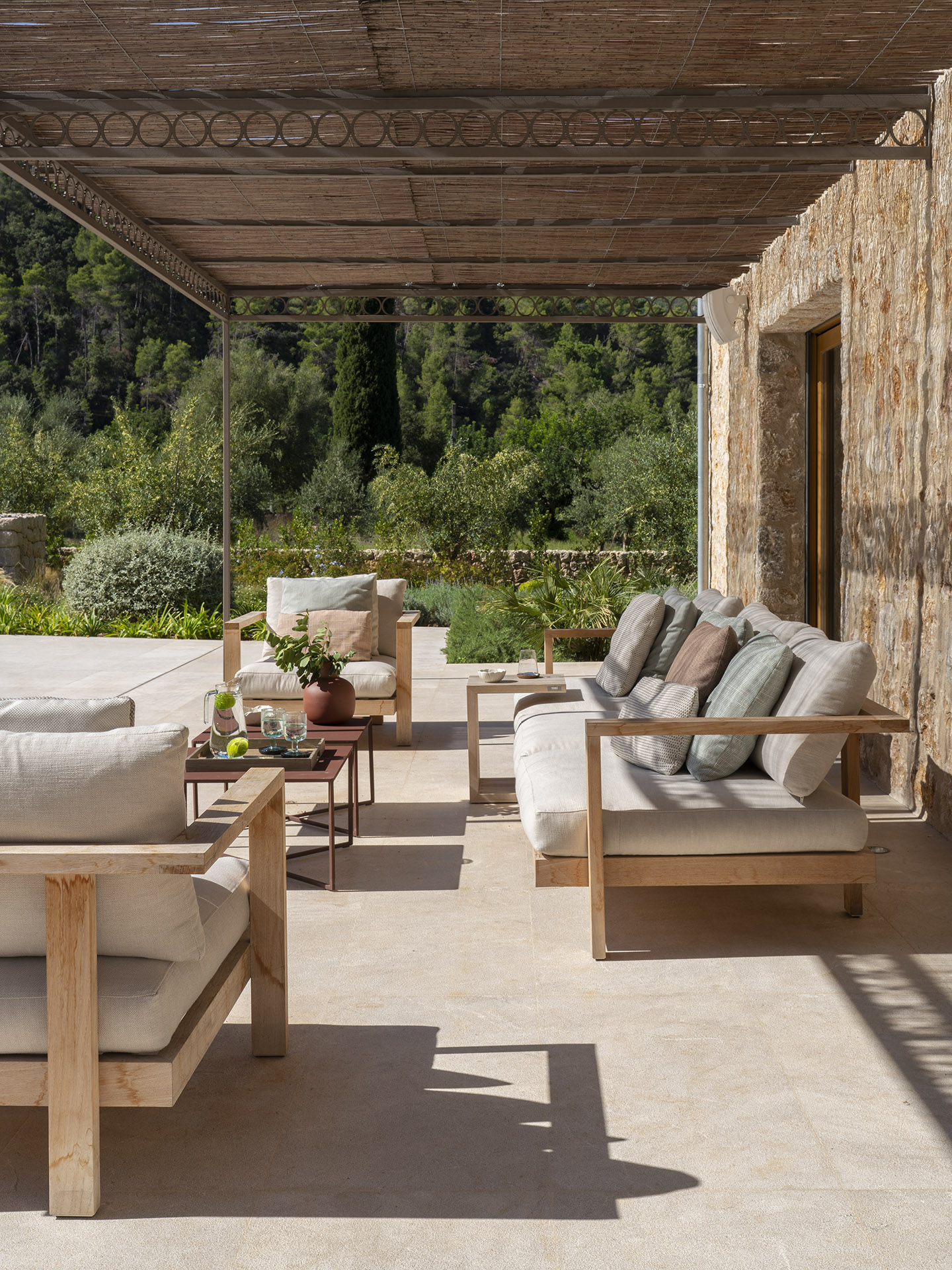 Terrasse mit Pergola aus Schilfrohr, Holzsofas und Holzsesseln, hellen Kissen und Steinwand
