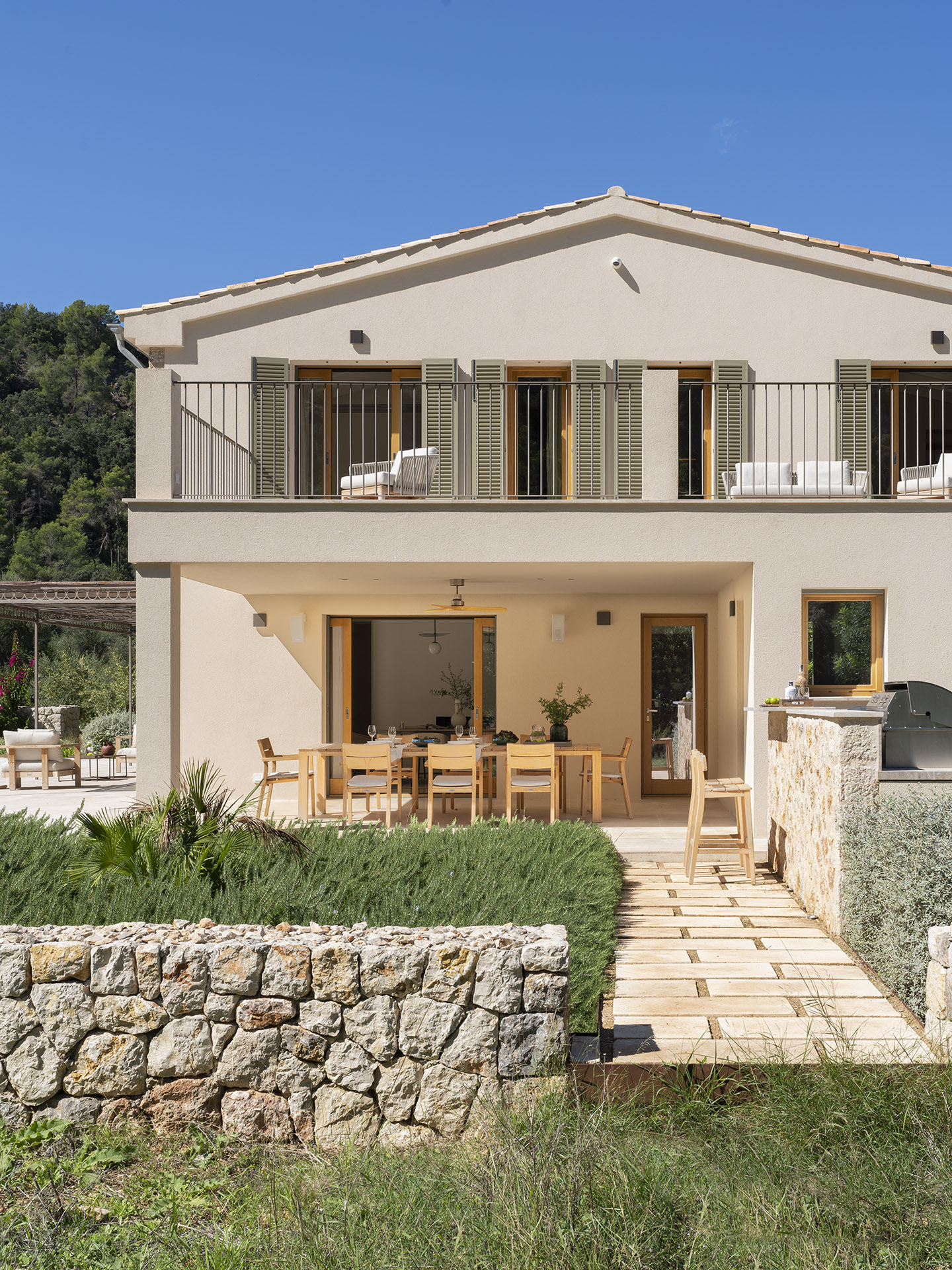 Zweistöckige Fassade mit Balkon, grünen Fensterläden, überdachtem Essbereich im Freien und Grill neben der Steinmauer