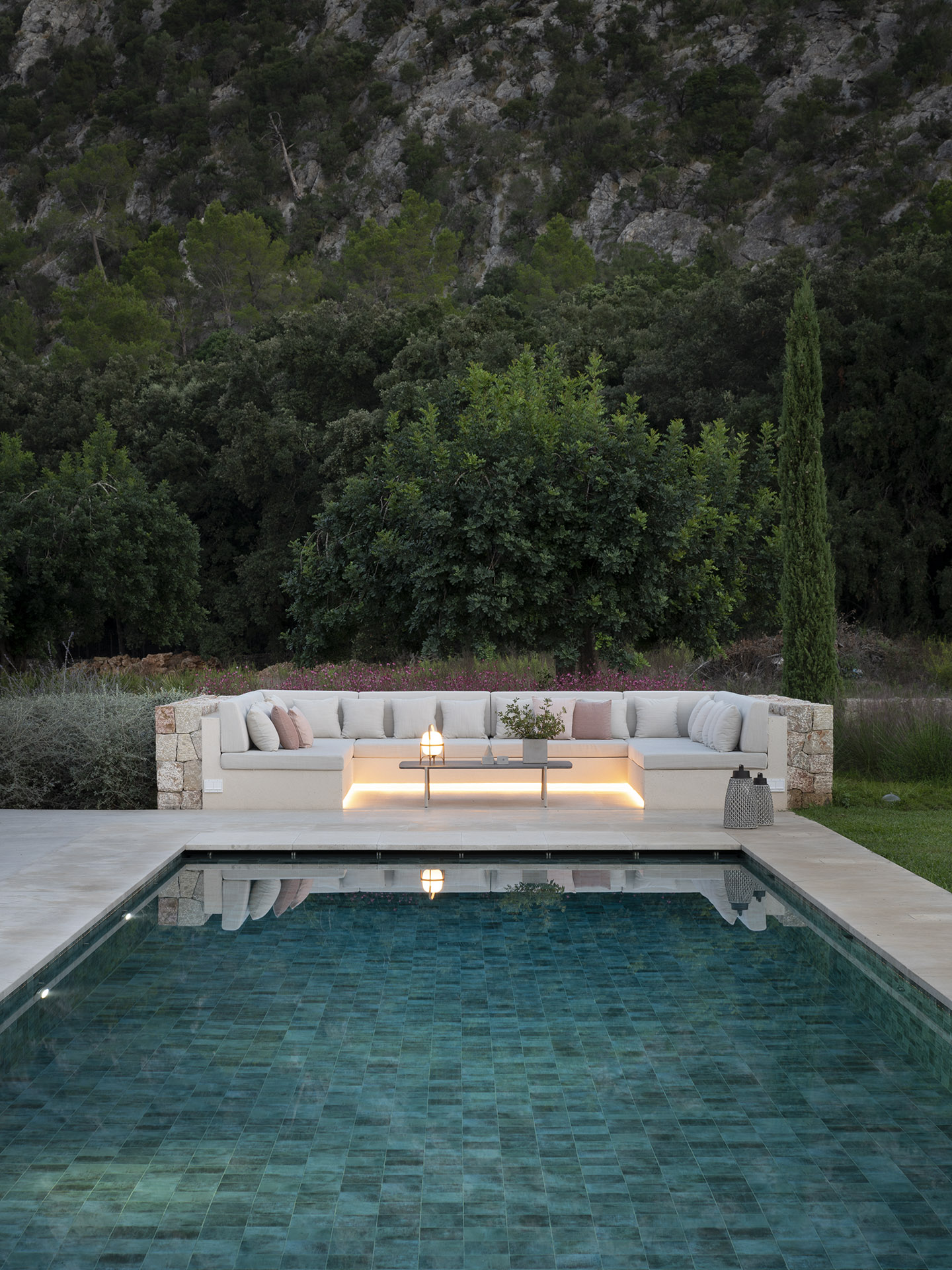 Chill-out-Bereich am Swimmingpool mit beleuchtetem gemauertem Sofa in der Abenddämmerung und bewaldetem Hang im Hintergrund