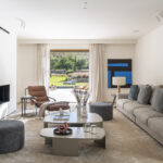 Geräumiges Wohnzimmer mit grauem Sofa, Ledersessel, blauem Gemälde und großer Fensterfront zum Garten und zum Swimmingpool