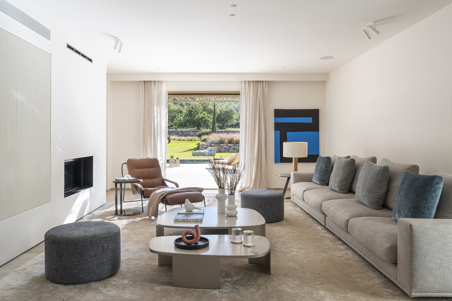 Geräumiges Wohnzimmer mit grauem Sofa, Ledersessel, blauem Gemälde und großer Fensterfront zum Garten und zum Swimmingpool