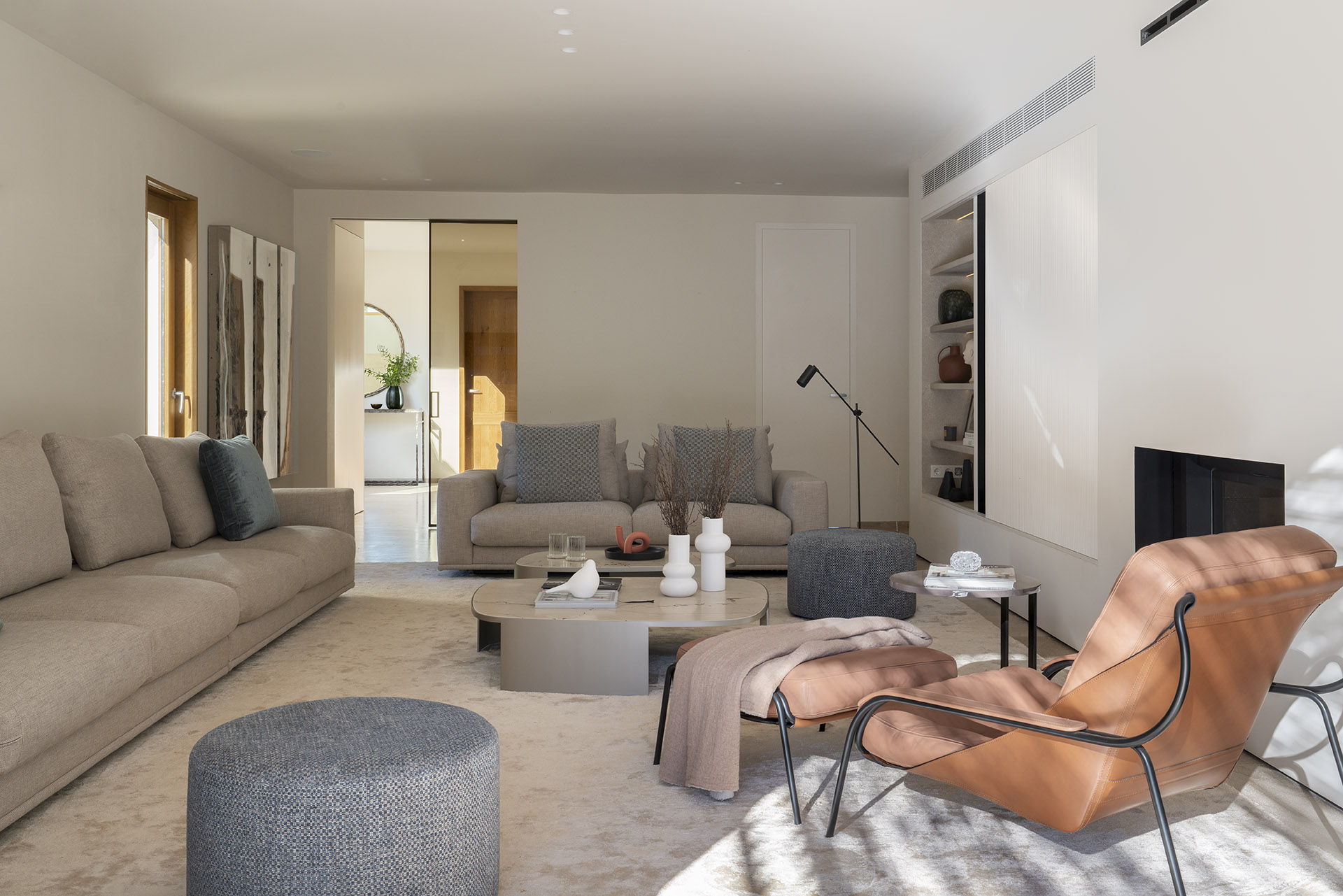 Wohnzimmer mit beigefarbenen Sofas, Ledersessel in natürlichem Lederton, grauen Poufs und Kamin rechts
