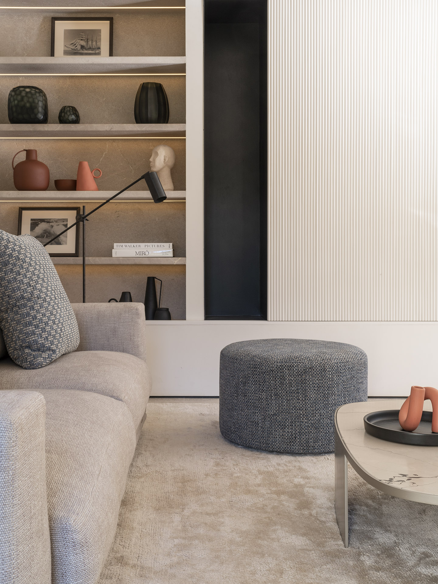 Detailansicht eines beleuchteten Regals mit Vasen und Büchern neben geriffeltem Paneel, grauem Pouf und Sofa