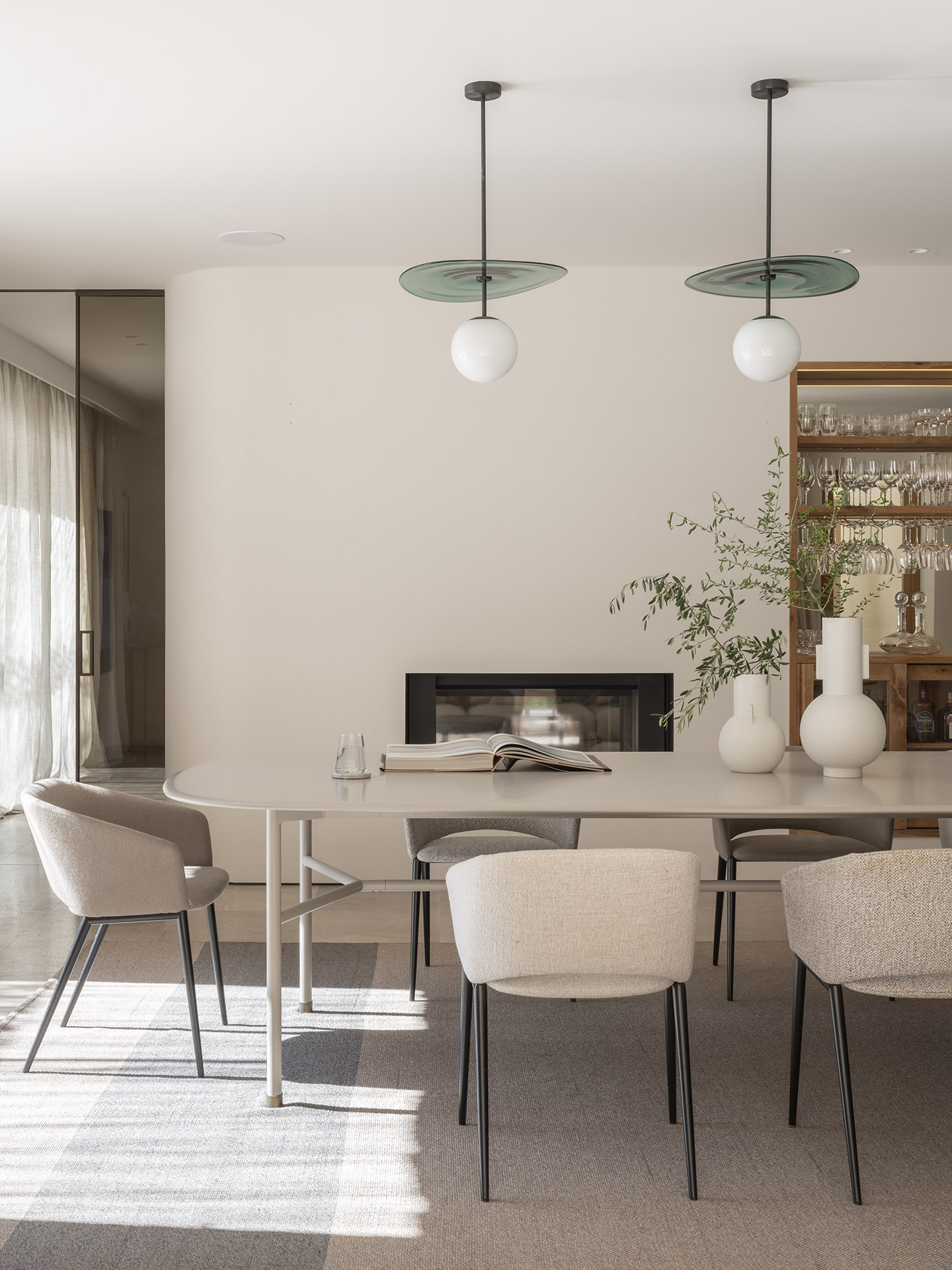 Esszimmer mit hellem ovalem Tisch, gepolsterten Stühlen, Hängeleuchten aus grünem Glas und Kamin im Hintergrund