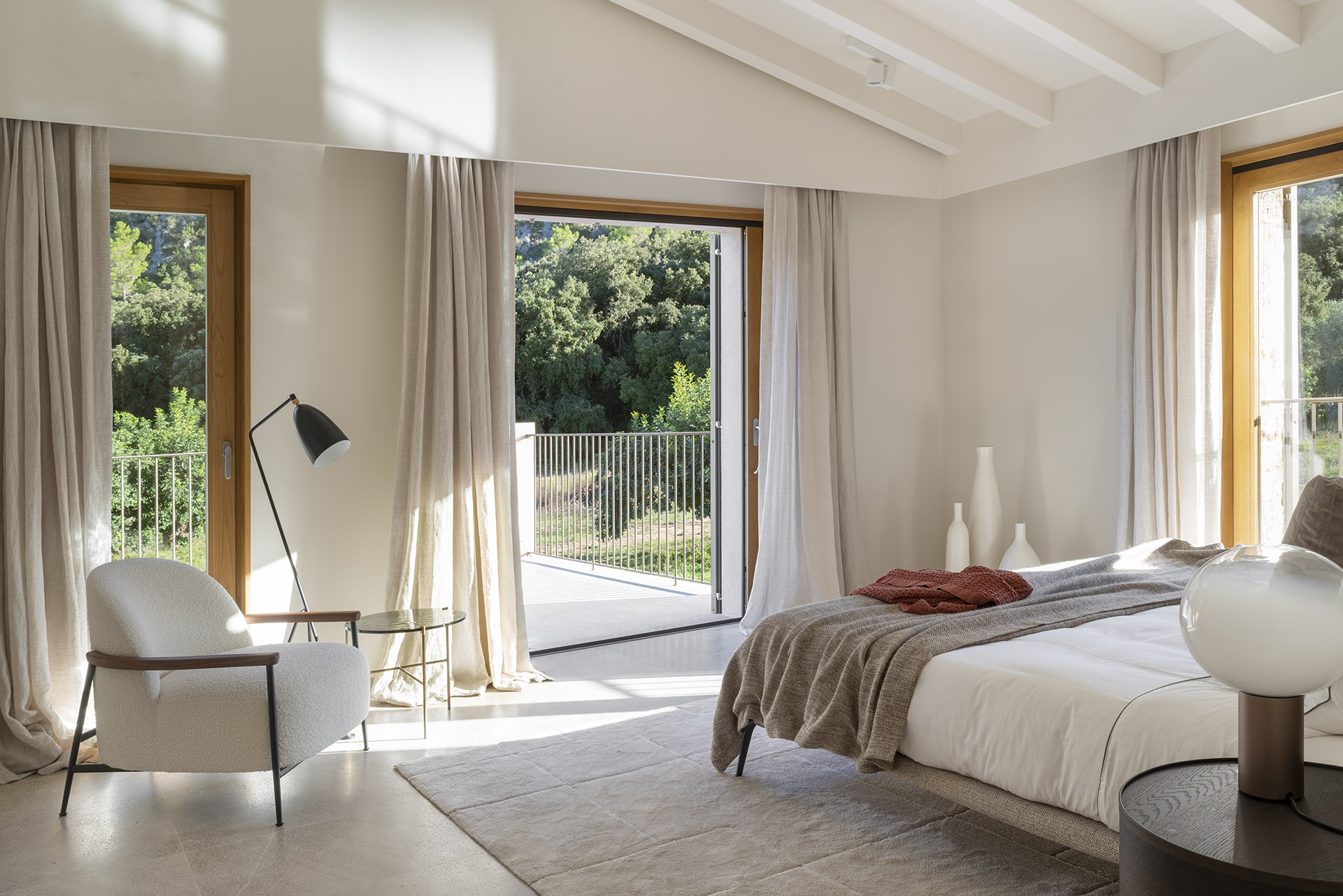 Schlafzimmer mit Leinenvorhängen und offener Holztür zu einer Terrasse mit Blick auf die Bäume