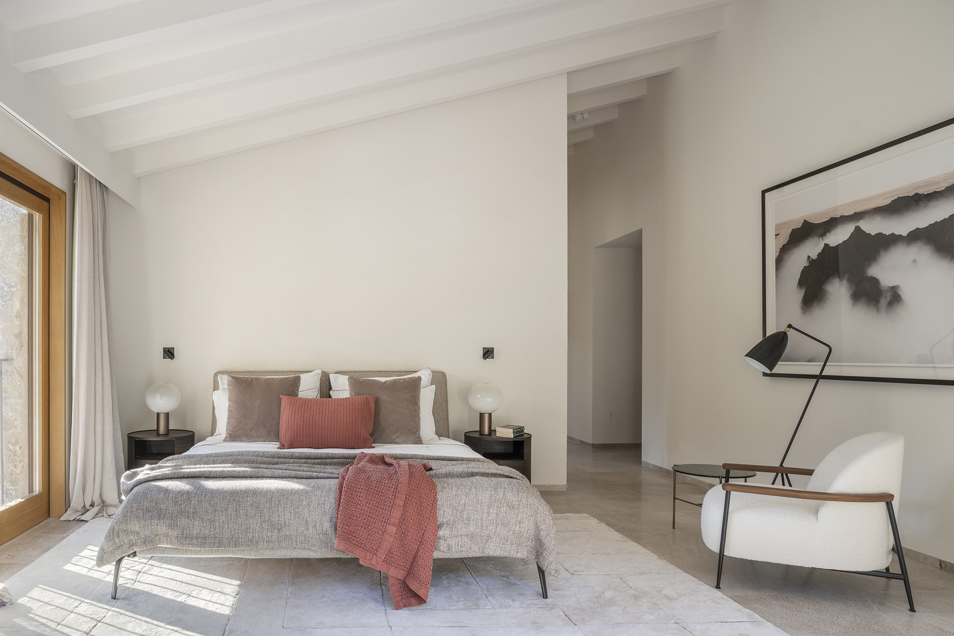 Schlafzimmer mit weißen Deckenbalken, Bett mit korallenroter Decke, weißem Sessel und schwarzer Stehlampe