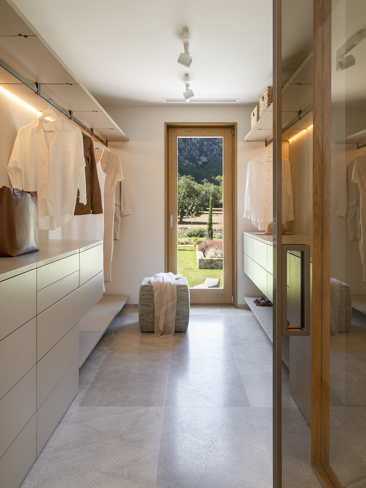Ankleidezimmer mit beigefarbenen Schränken, aufgehängten Hemden, Holztür zum Garten und hellem Steinboden