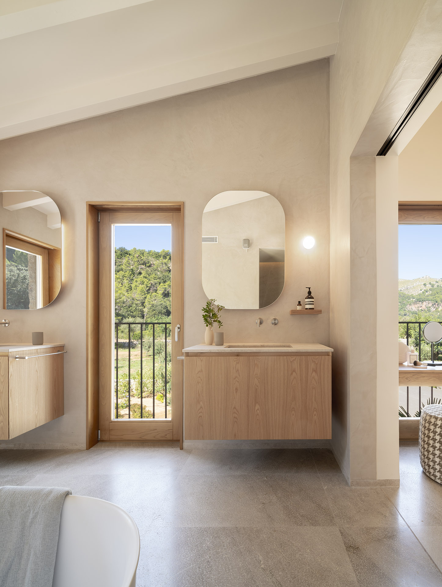 Badezimmer mit abgerundeten Spiegeln, wandhängenden Holzwaschtischen und Balkontür mit Blick auf die Bäume