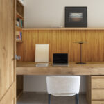 Büro mit Holzschreibtisch, beleuchteter Wand aus senkrechten Lamellen, weißem Stuhl und Regal mit Büchern