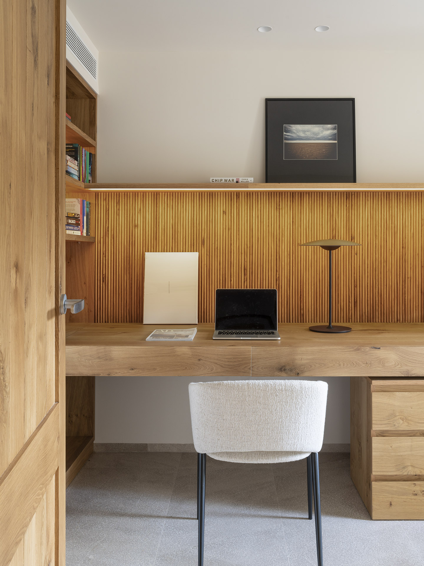 Büro mit Holzschreibtisch, beleuchteter Wand aus senkrechten Lamellen, weißem Stuhl und Regal mit Büchern