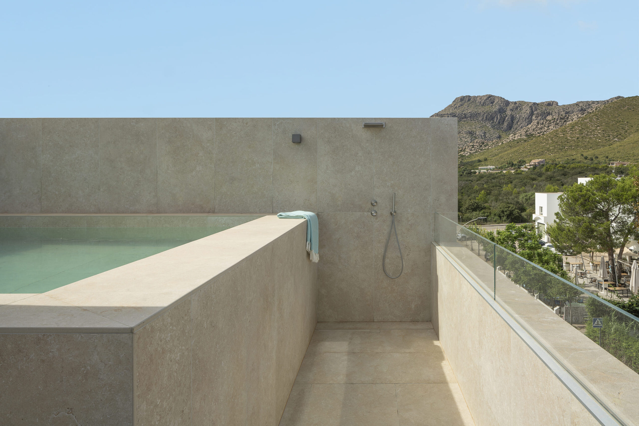 Swimmingpool und Dusche auf der Dachterrasse, mit beigem Stein verkleidet, dazu Glasgeländer und Blick auf die Berge