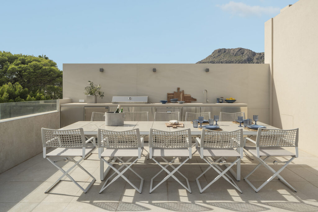 Essbereich im Freien auf der Dachterrasse mit langem Tisch, weißen Stühlen, Grill und Bergen im Hintergrund