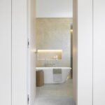 Bad mit weißer freistehender Badewanne, beiger Steinverkleidung und beleuchteter Nische, Durchblick zwischen weißen Schränken