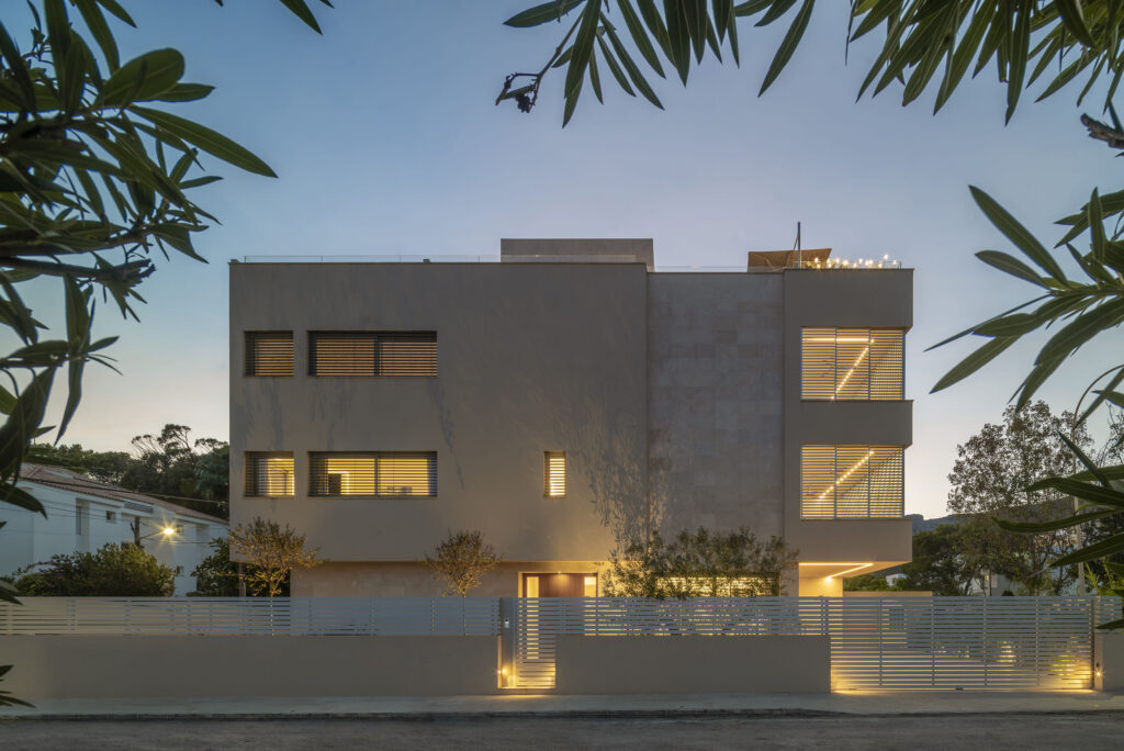 Fachada de la casa Far al anochecer con ventanas iluminadas y valla de lamas blancas