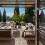 Covered terrace seen through a glazed door, with wooden sofas, coffee table, cypress trees and reflections