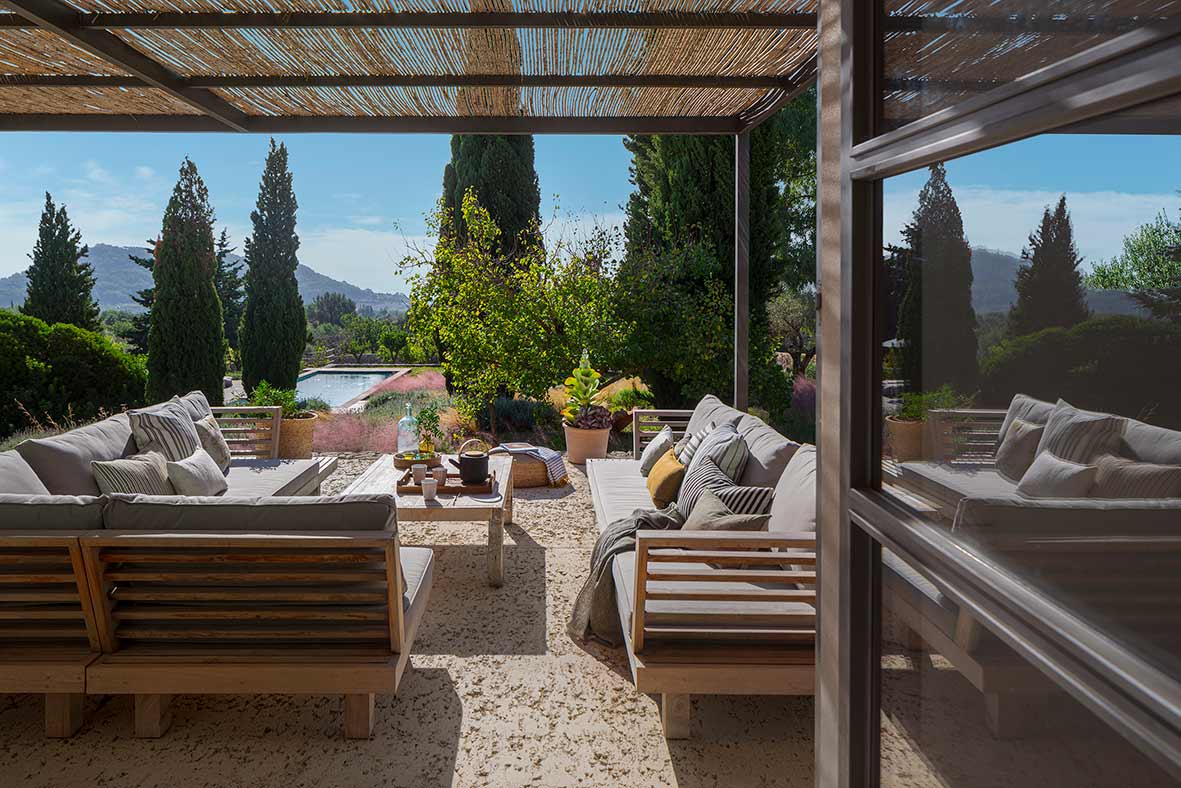 Outdoor porch with wooden sofas and grey cushions, reed pergola, garden, pool and mountains beyond