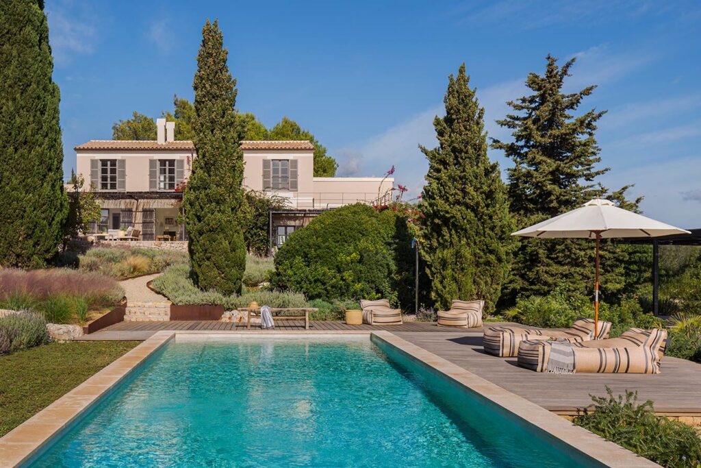 Finca Rosa seen from the pool, with sun loungers under a parasol, cypress trees and a pale-toned façade
