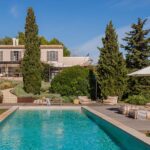 Finca Rosa seen from the pool, with sun loungers under a parasol, cypress trees and a pale-toned façade
