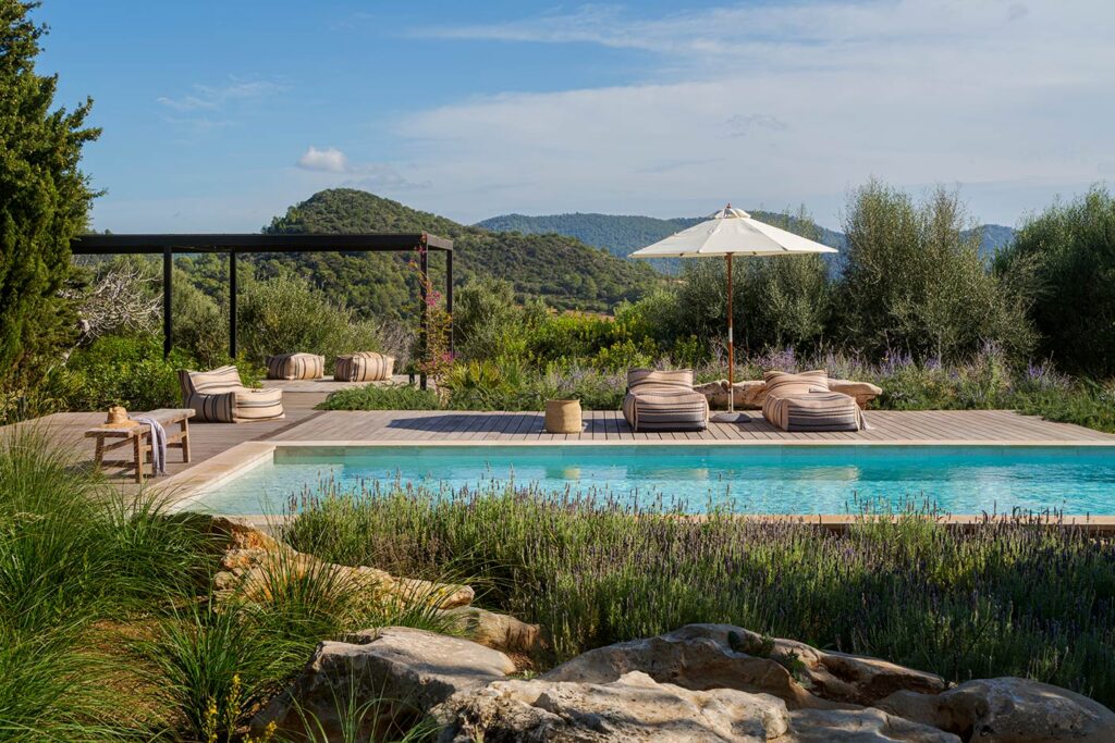 Pool with wooden sun deck, striped pouffes and parasol, surrounded by lavender, rocks and wooded mountains