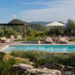 Pool with wooden sun deck, striped pouffes and parasol, surrounded by lavender, rocks and wooded mountains