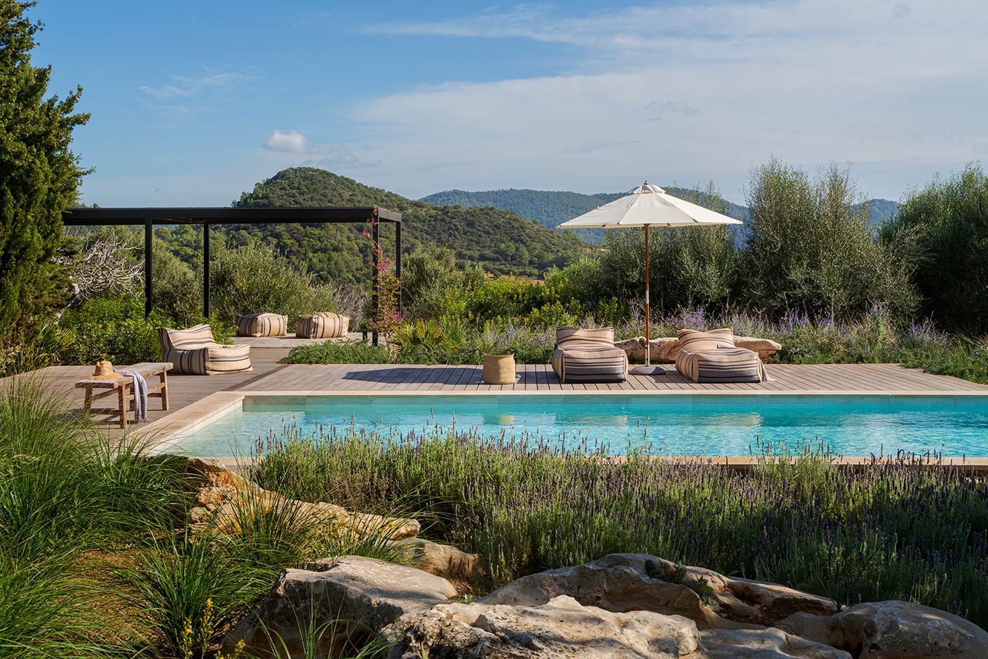 Pool with wooden sun deck, striped pouffes and parasol, surrounded by lavender, rocks and wooded mountains