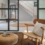 Jute pouffe and wooden armchair in the living room, with white staircase beyond a glazed screen