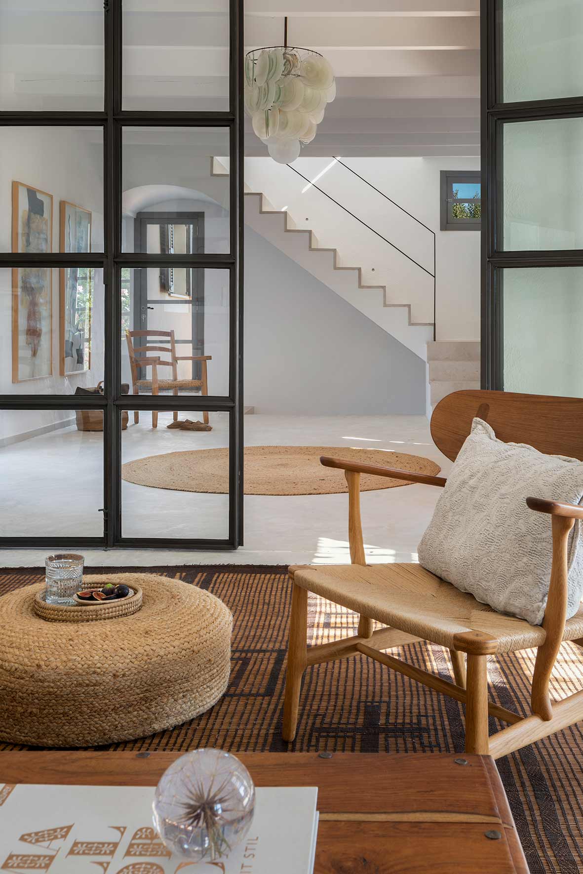 Jute pouffe and wooden armchair in the living room, with white staircase beyond a glazed screen