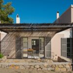 Façade with reed-covered metal pergola, grey shutters, stone terrace, rubble-stone wall and metal chairs
