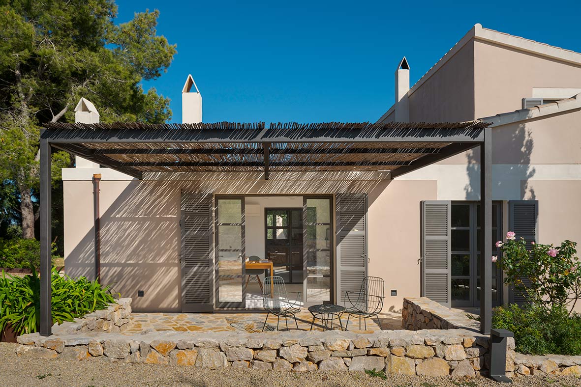 Façade with reed-covered metal pergola, grey shutters, stone terrace, rubble-stone wall and metal chairs